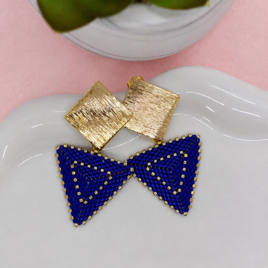 Blue triangle earrings on a white dish with a gold square earring and decorative plate in the background.