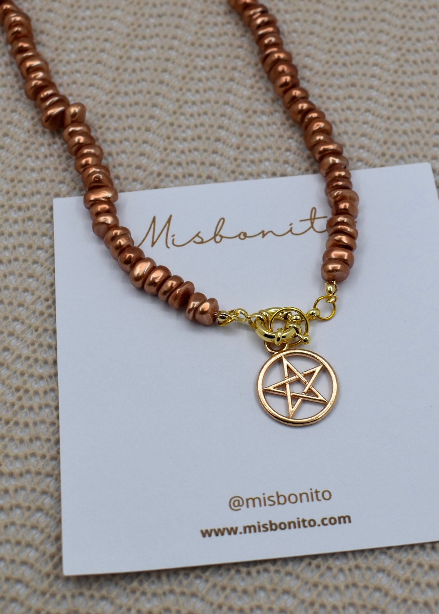 A copper-colored beaded necklace with a gold pentagram pendant, displayed on a white Misbonito brand card over a textured beige fabric background.