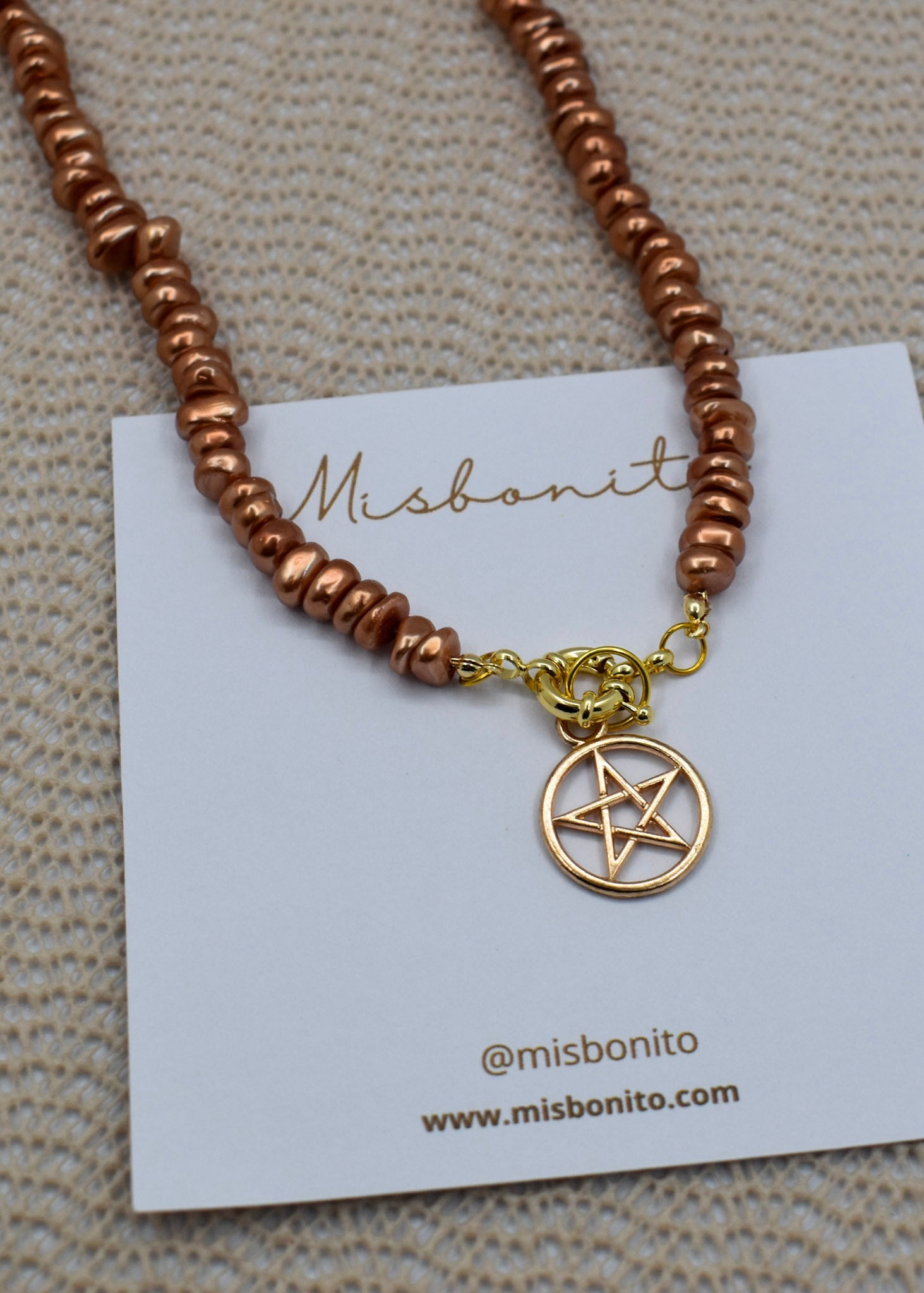 A copper-colored beaded necklace with a gold pentagram pendant, displayed on a white Misbonito brand card over a textured beige fabric background.