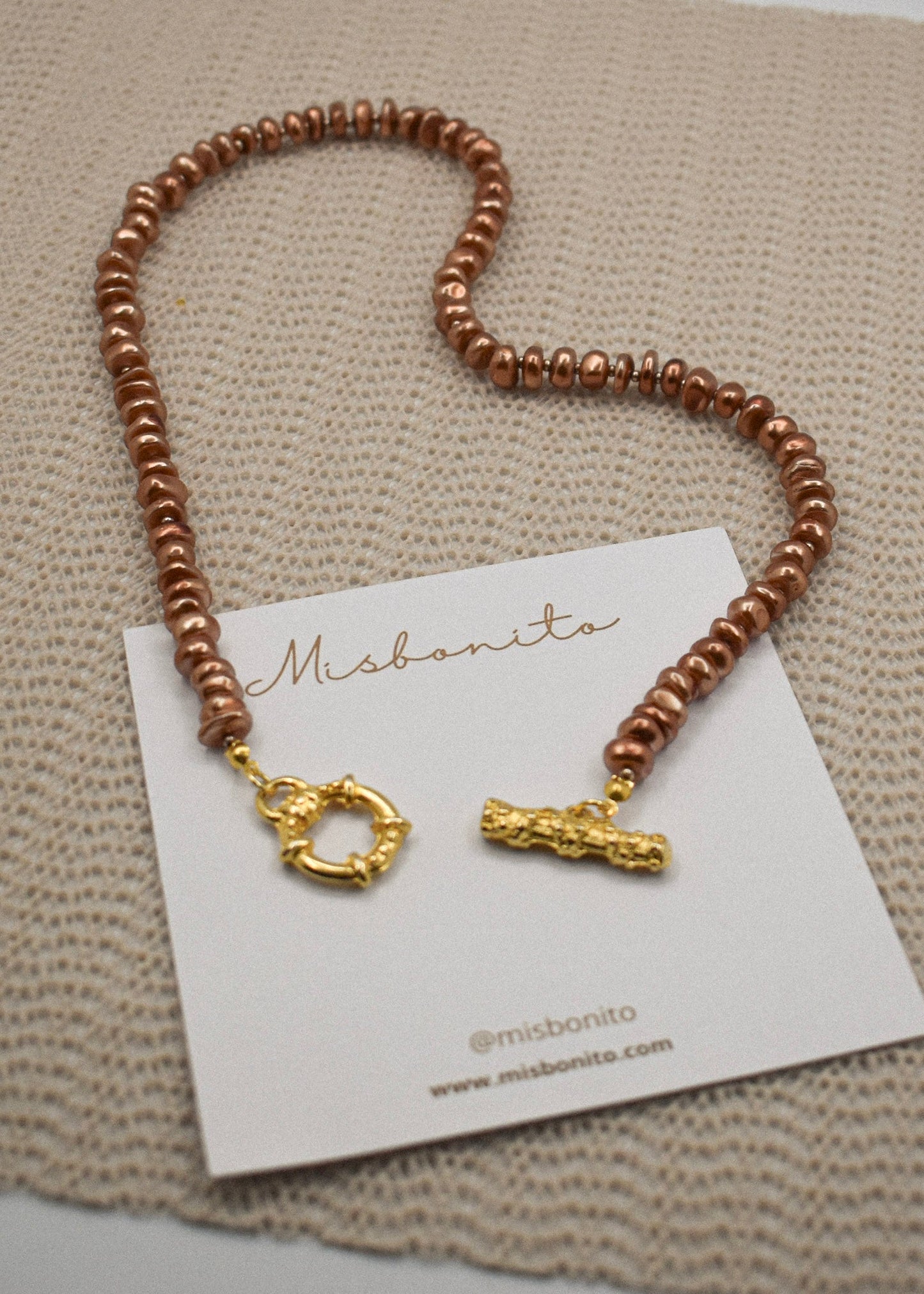 A copper-colored beaded necklace with gold-toned hardware, displayed on a white Misbonito brand card over a textured beige woven fabric background.