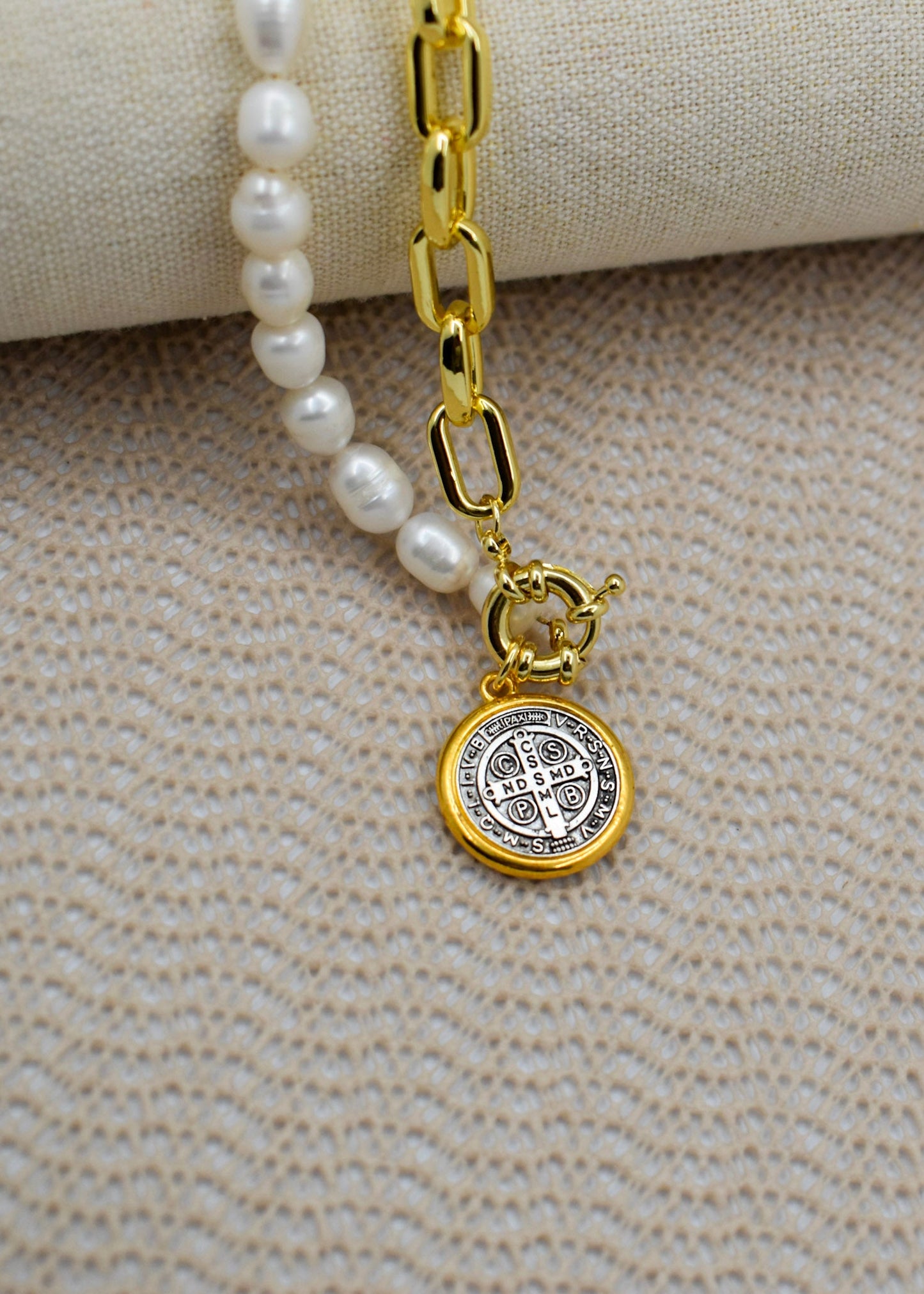A close-up of a necklace with white pearl-like beads, a gold chain link section, and a round gold Saint Benedict medallion pendant, displayed on a textured beige fabric surface.