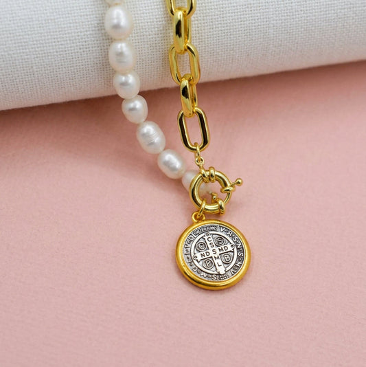 A close-up of a necklace with white pearl-like beads, a gold chain link section, and a round gold Saint Benedict medallion pendant, displayed on a textured beige fabric surface.