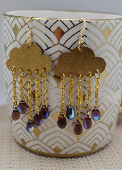 A pair of gold cloud-shaped dangle earrings with iridescent teardrop beads, hanging from a white ceramic mug with a gold handle and geometric pattern.