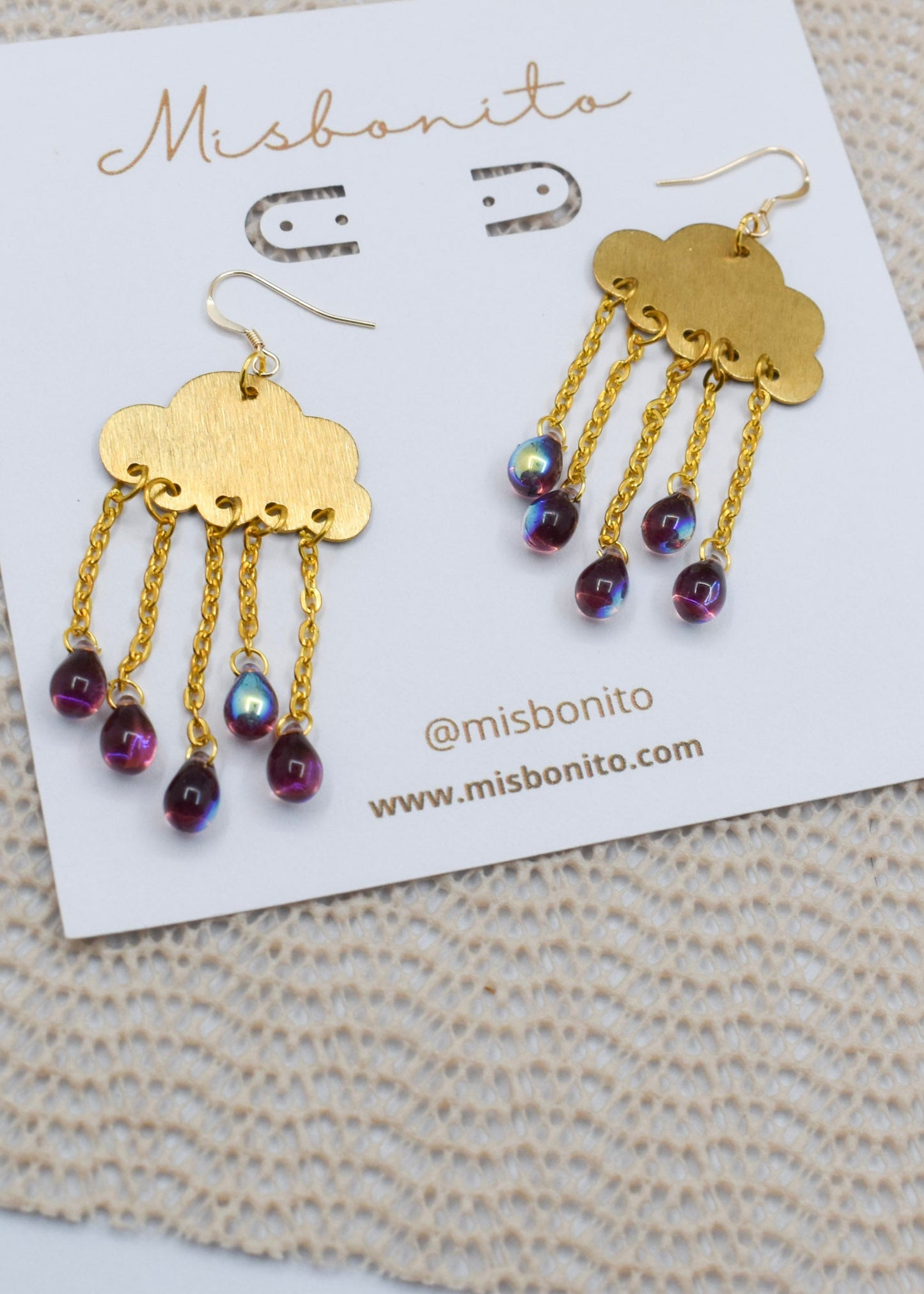 A pair of Misbonito cloud-shaped gold dangle earrings with iridescent purple beads, displayed on a white branded card over a beige knit fabric background.