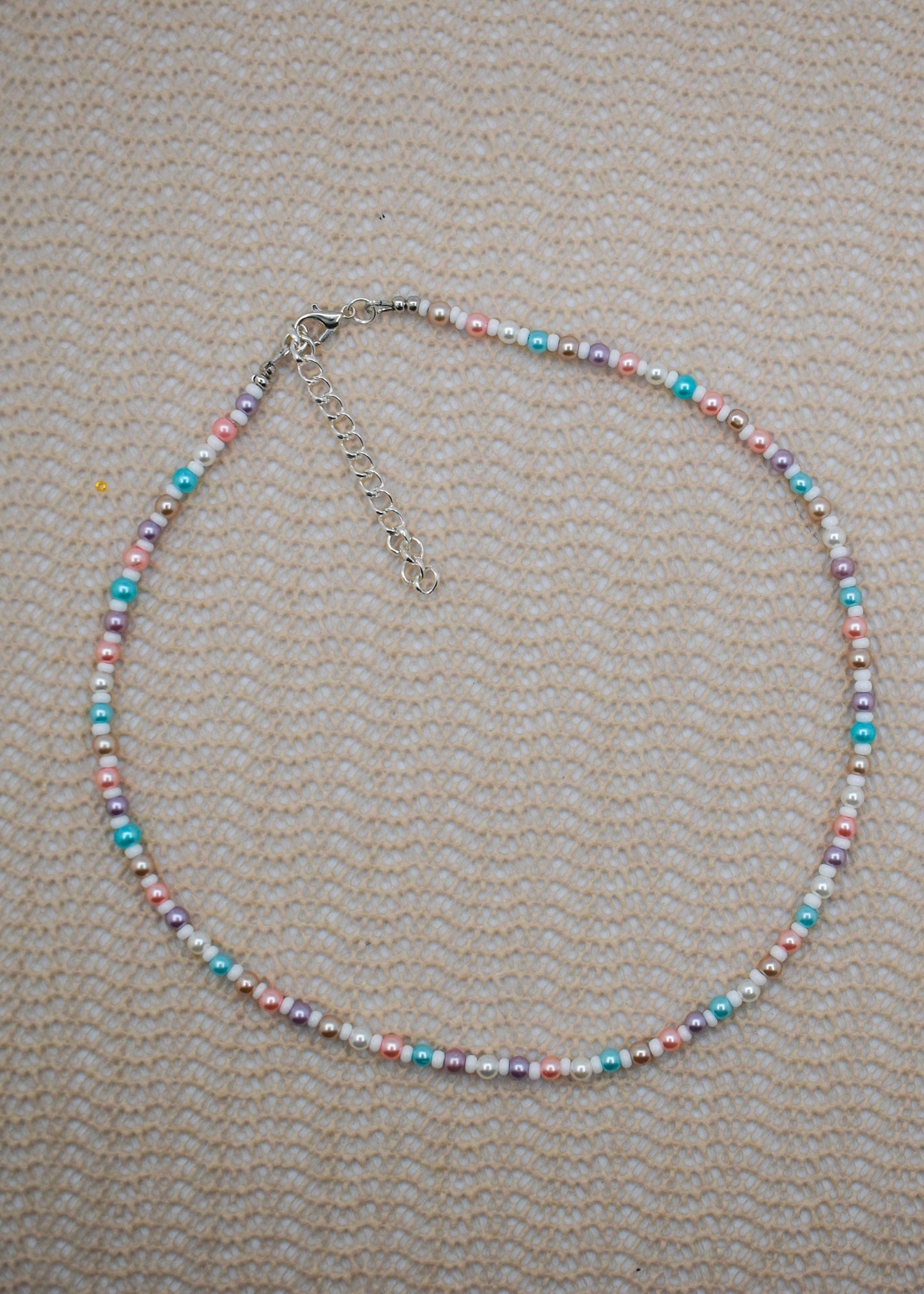 A beaded necklace with small pastel-colored beads in pink, blue, purple, and white, accented by silver-toned spacer beads and a silver clasp with extender chain, laid flat on a textured beige fabric background.