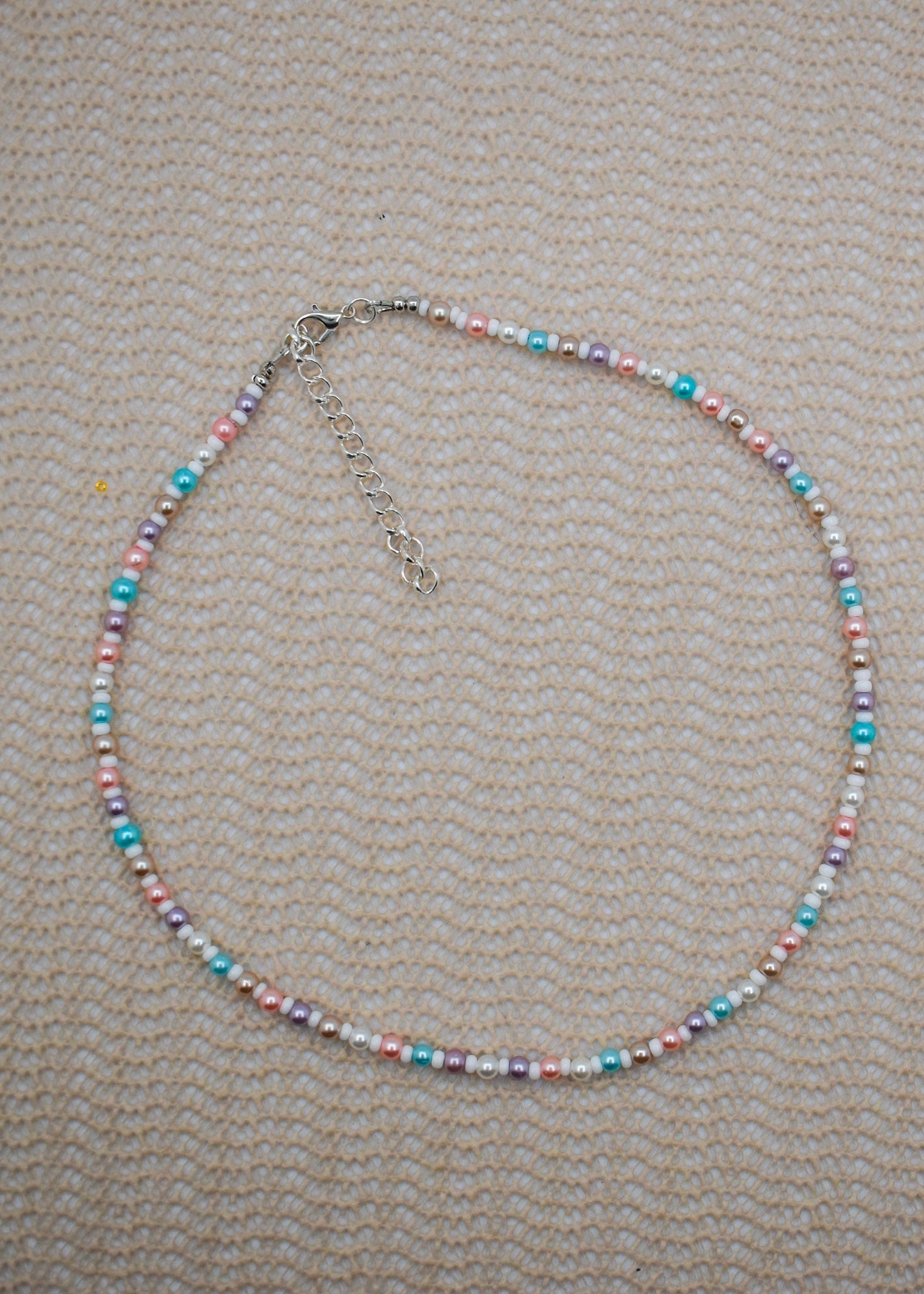 A beaded necklace with small pastel-colored beads in pink, blue, purple, and white, accented by silver-toned spacer beads and a silver clasp with extender chain, laid flat on a textured beige fabric background.