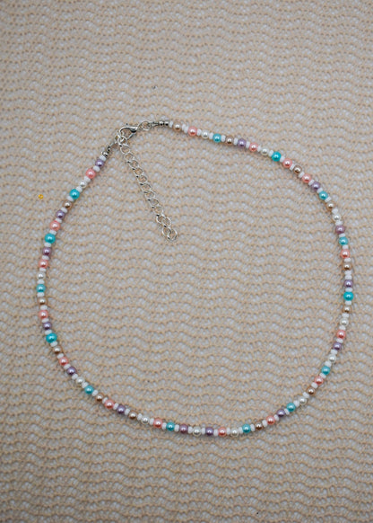 A beaded necklace with small pastel-colored beads in pink, blue, purple, and white, accented by silver-toned spacer beads and a silver clasp with extender chain, laid flat on a textured beige fabric background.