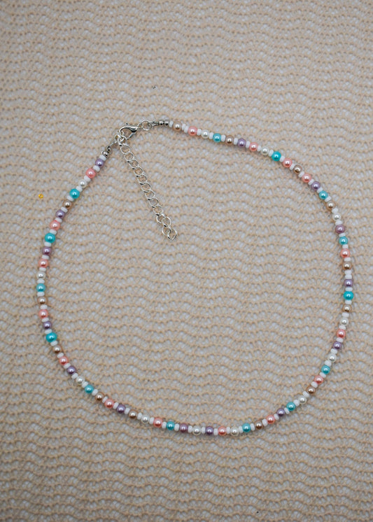 A beaded necklace with small pastel-colored beads in pink, blue, purple, and white, accented by silver-toned spacer beads and a silver clasp with extender chain, laid flat on a textured beige fabric background.