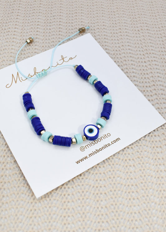 A beaded bracelet with dark blue cylindrical beads, light blue round beads, and a central blue and white evil eye charm, displayed on a white branded card on a textured beige surface.