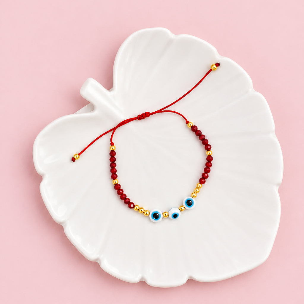 A beaded bracelet with red, gold, and blue evil eye beads, displayed on a white leaf-shaped ceramic dish over a beige textured fabric background.