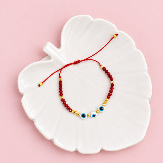 A beaded bracelet with red, gold, and blue evil eye beads, displayed on a white leaf-shaped ceramic dish over a beige textured fabric background.