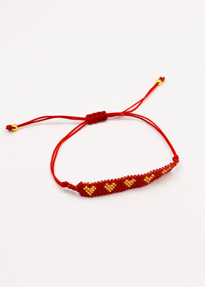A red cord bracelet with a beaded bar featuring golden and red geometric patterns, shown on a plain white background.