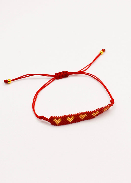 A red cord bracelet with a beaded bar featuring golden and red geometric patterns, shown on a plain white background.