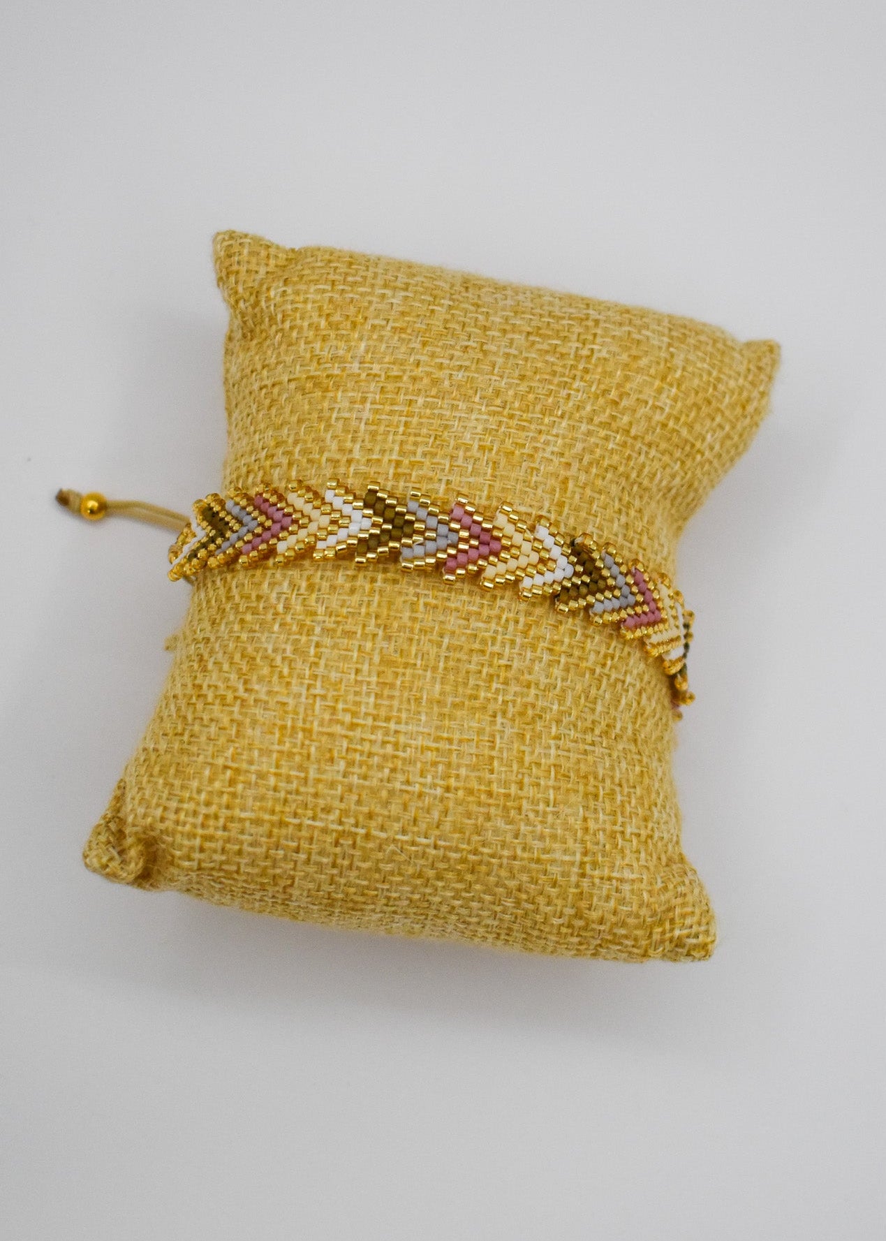 A gold beaded chevron bracelet with red and white accents, displayed on a small yellow fabric jewelry pillow against a white background.