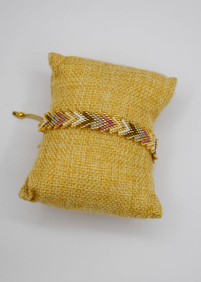 A gold beaded chevron bracelet with red and white accents, displayed on a small yellow fabric jewelry pillow against a white background.