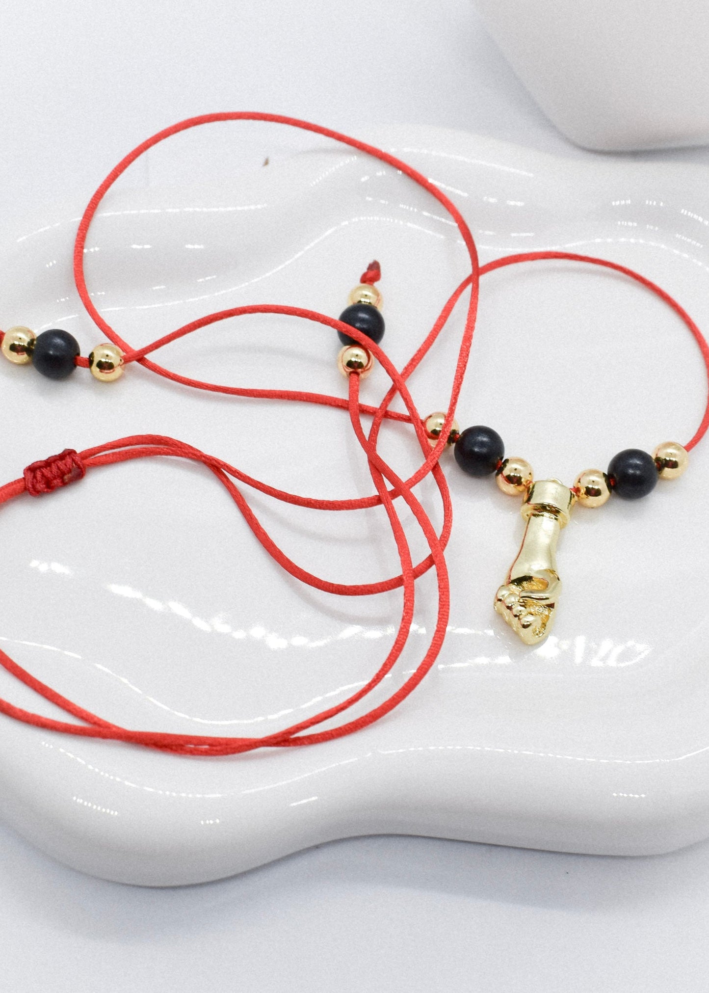 Red cord bracelet with black and gold beads and a hand charm on a white dish