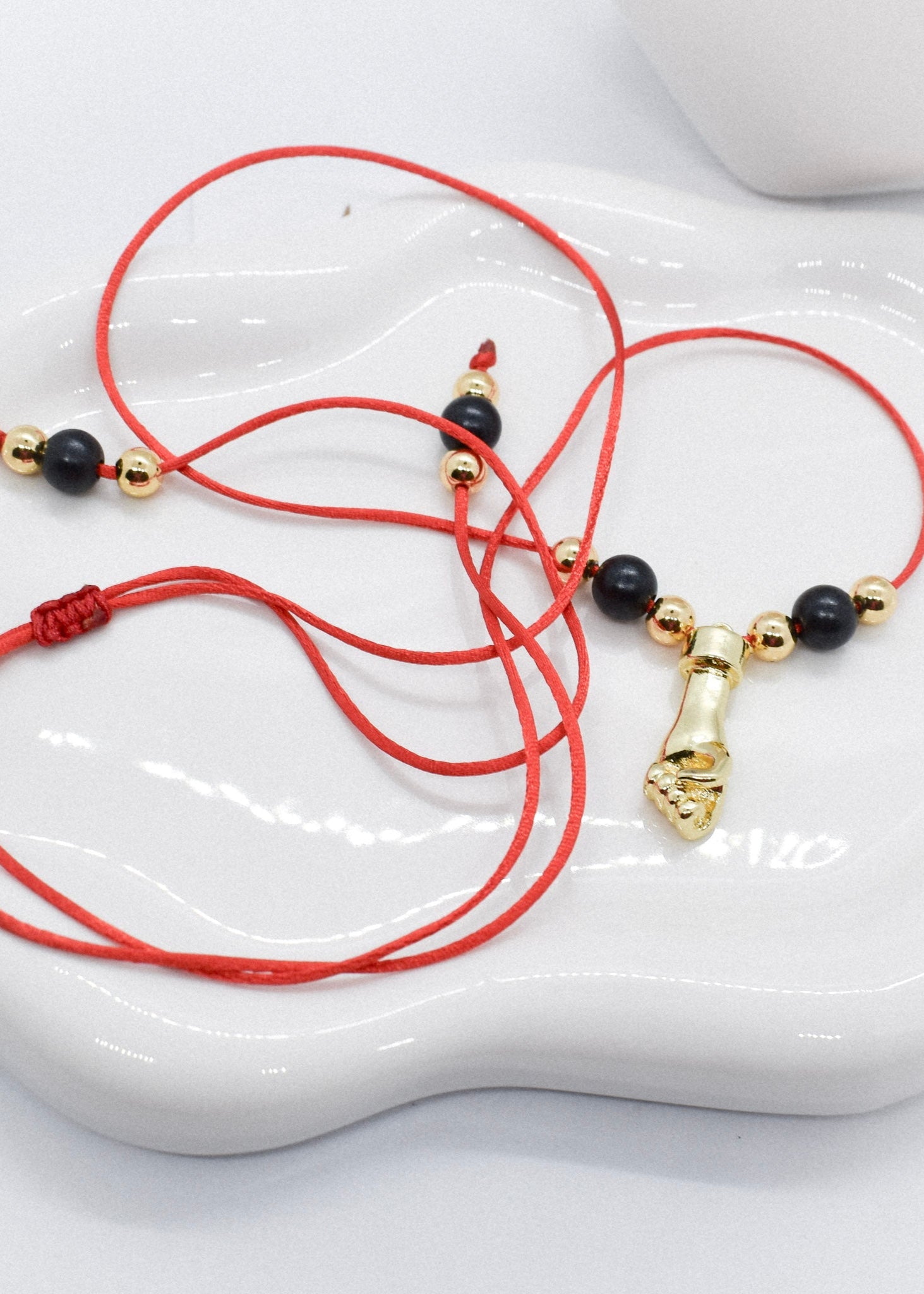Red cord bracelet with black and gold beads and a hand charm on a white dish