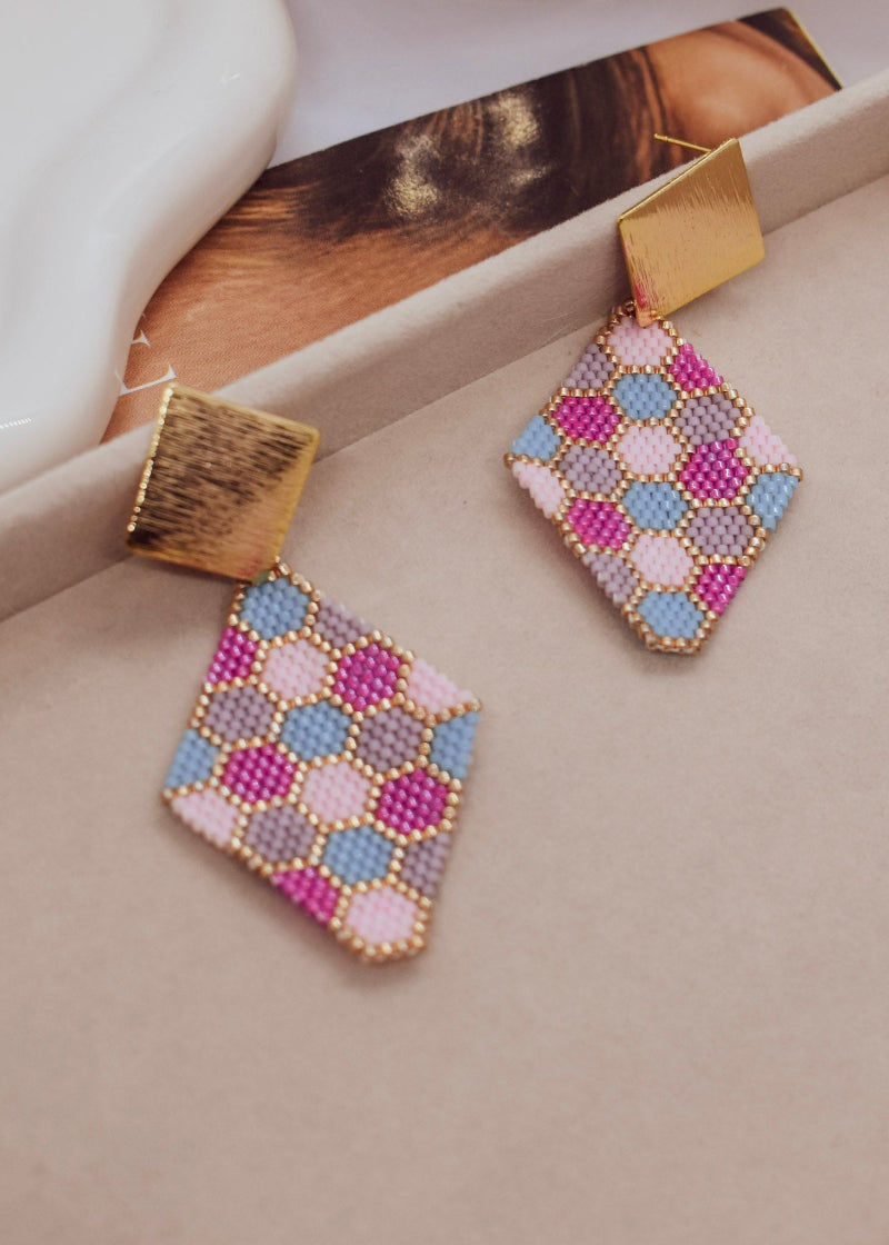 A pair of handmade geometric dangle earrings with gold-tone metal tops and diamond-shaped beaded pendants featuring a pink, blue, and gray geometric pattern, displayed on a neutral beige surface.
