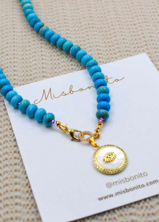 A jasper beaded necklace with a gold-toned clasp and a round white pendant featuring a gold eye design, displayed on a white card with the brand name Misbonito.