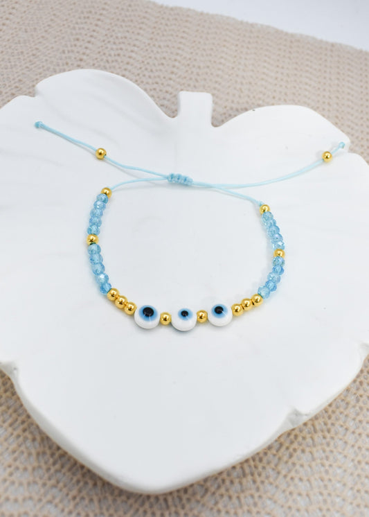 A light blue beaded bracelet with three white evil eye charms and gold spacer beads, displayed on a white ceramic heart-shaped dish over a beige textured fabric background.