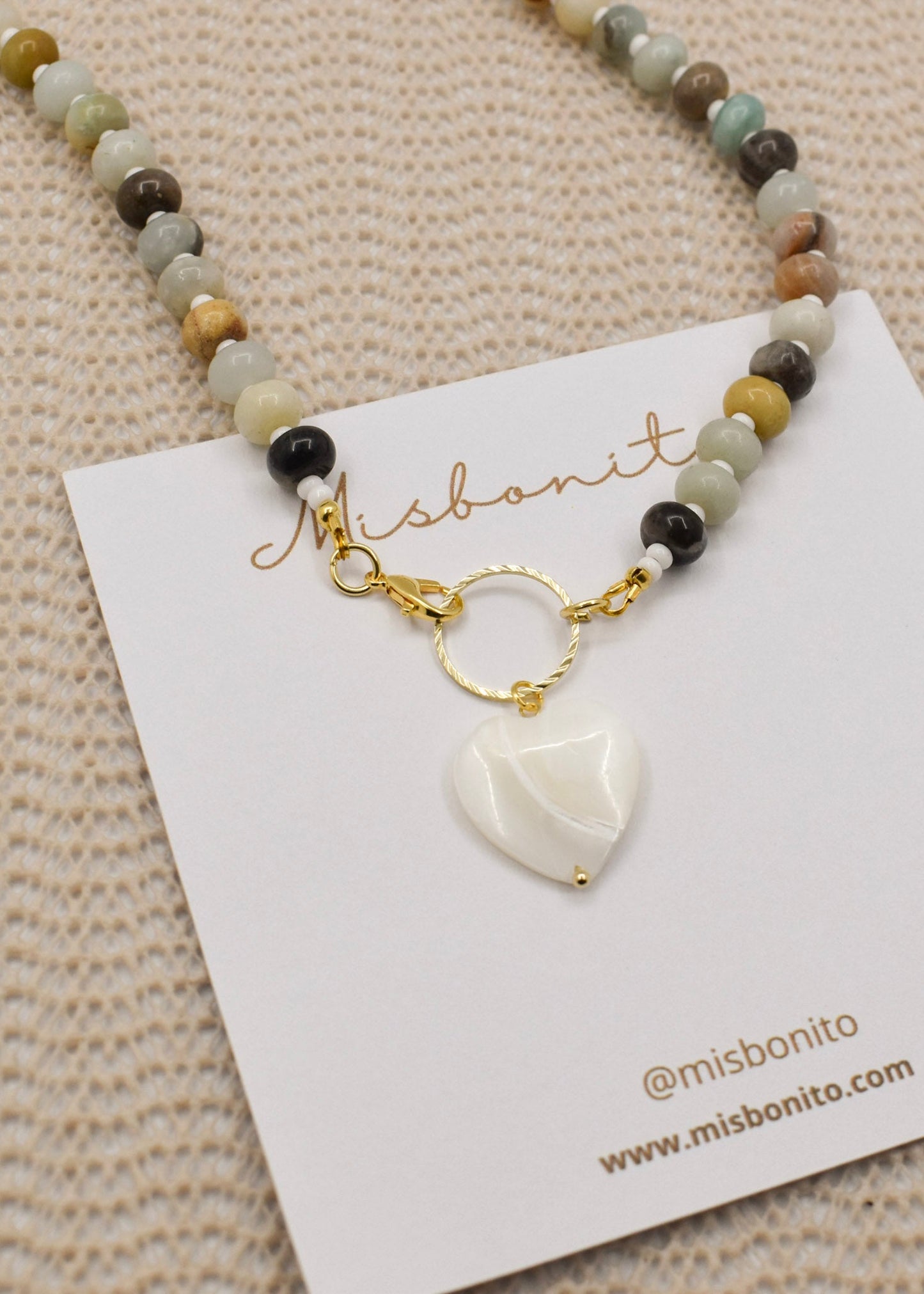 A beaded necklace with a white heart-shaped pendant and gold clasp, displayed on a white card labeled Misbonito over a beige textured fabric background.