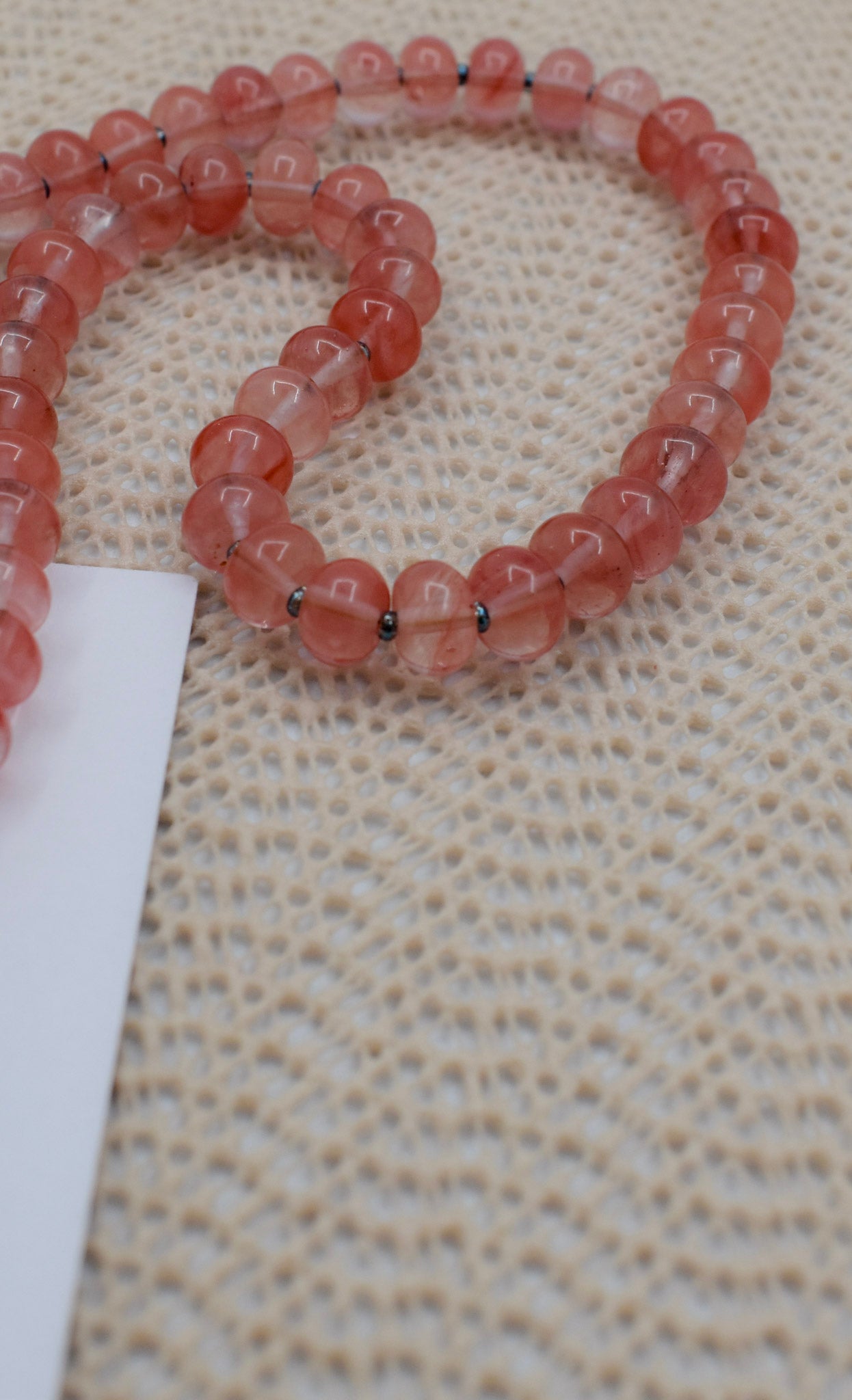 A strand of translucent pinkish-red round beads with small dark spacer rings, laid on a textured beige woven surface next to a white card.