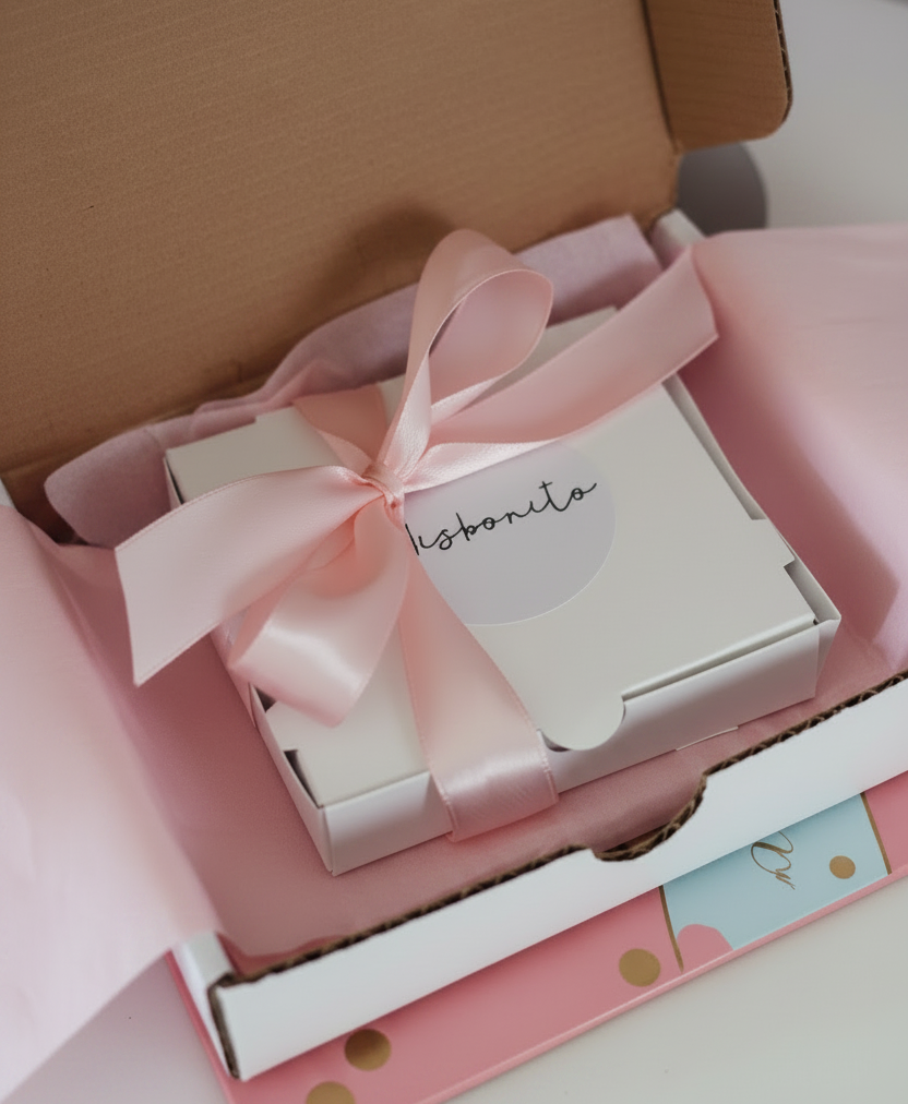 A small white gift box tied with a pale pink satin ribbon and bow, placed inside an open brown cardboard box lined with pink tissue paper. The top of the white box shows the script brand name 'lisbona'.