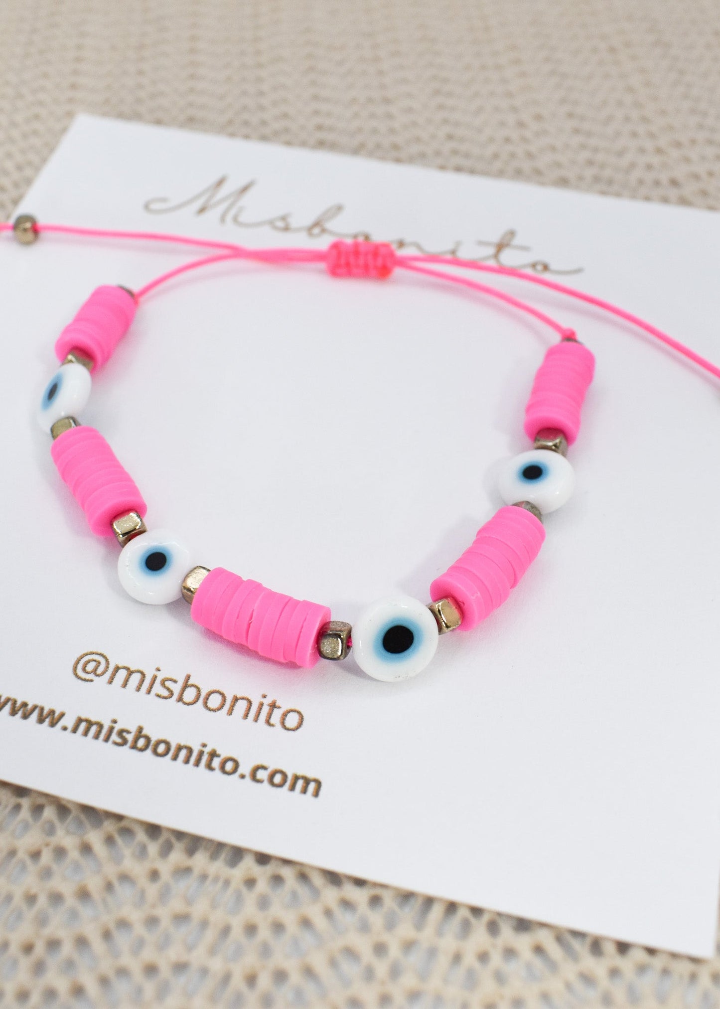 A pink cord bracelet with pink cylindrical beads, white evil eye beads, and small metallic spacer beads, displayed on a white Misbonito card.
