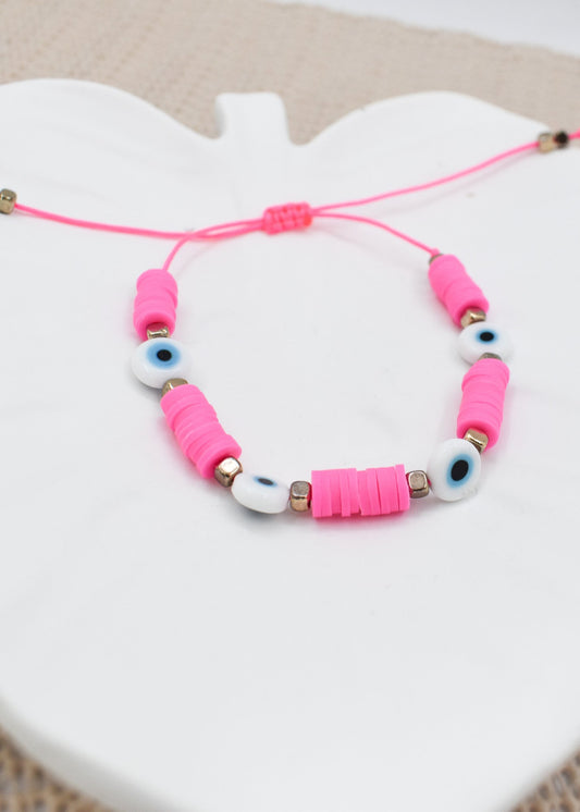 A pink cord bracelet with pink wrapped beads, white evil eye beads, and small gold spacer beads, displayed on a white ceramic shell-shaped plate.