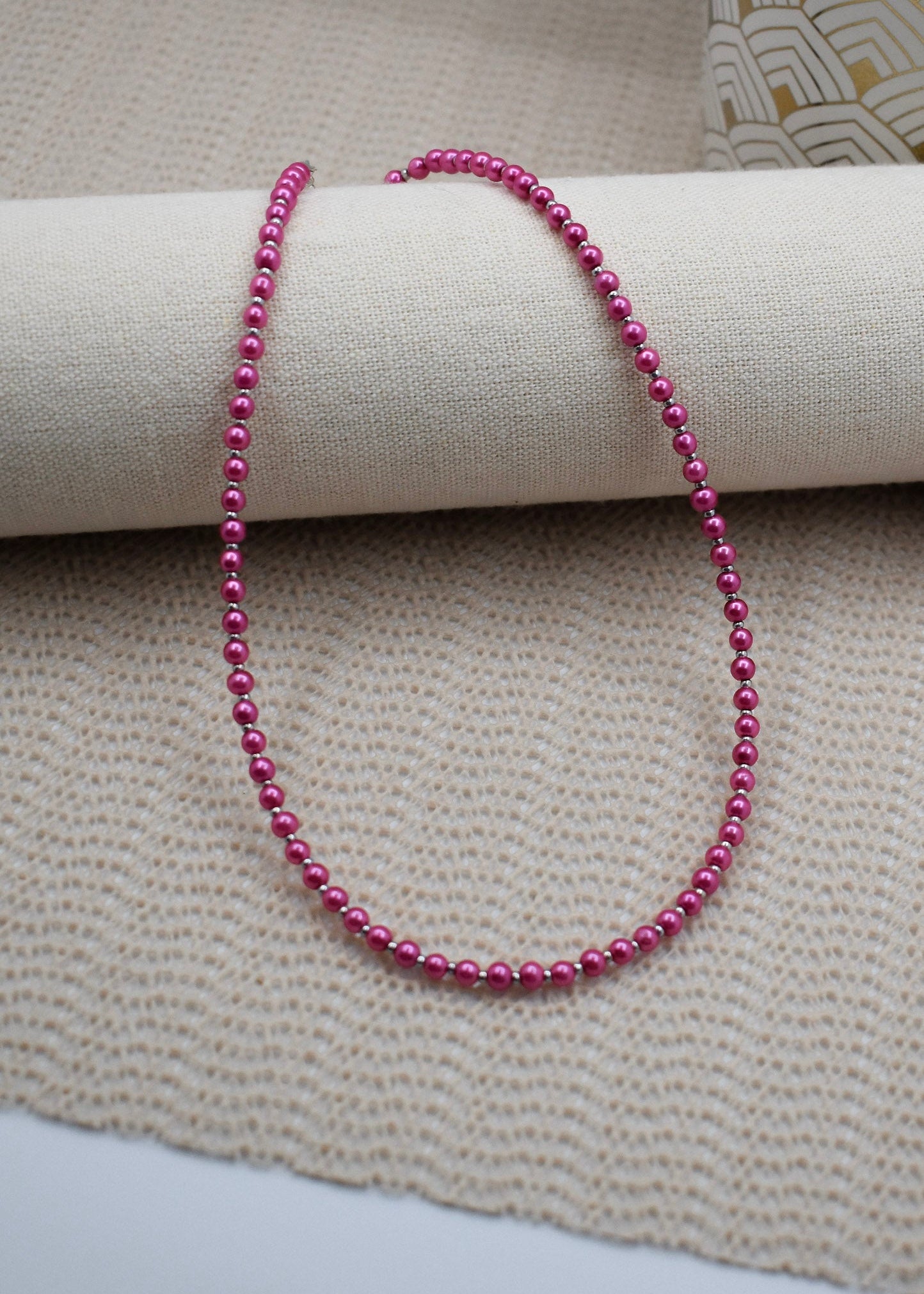 A strand of small, glossy magenta-pink beads draped over a cream-colored fabric roll, set against a textured beige woven fabric background.