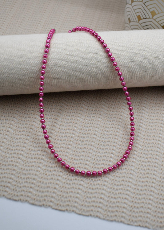 A strand of small, glossy magenta-pink beads draped over a cream-colored fabric roll, set against a textured beige woven fabric background.
