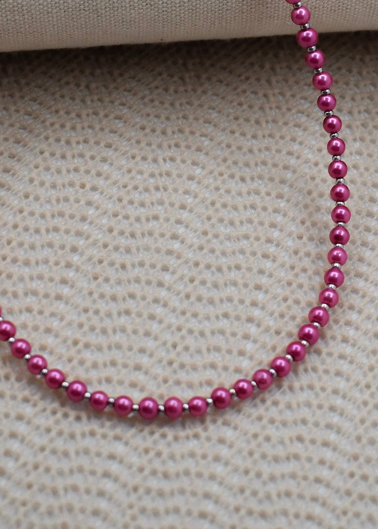 A close-up of a beaded necklace with glossy magenta-pink round beads separated by small metallic spacer rings, draped on a textured beige fabric background.
