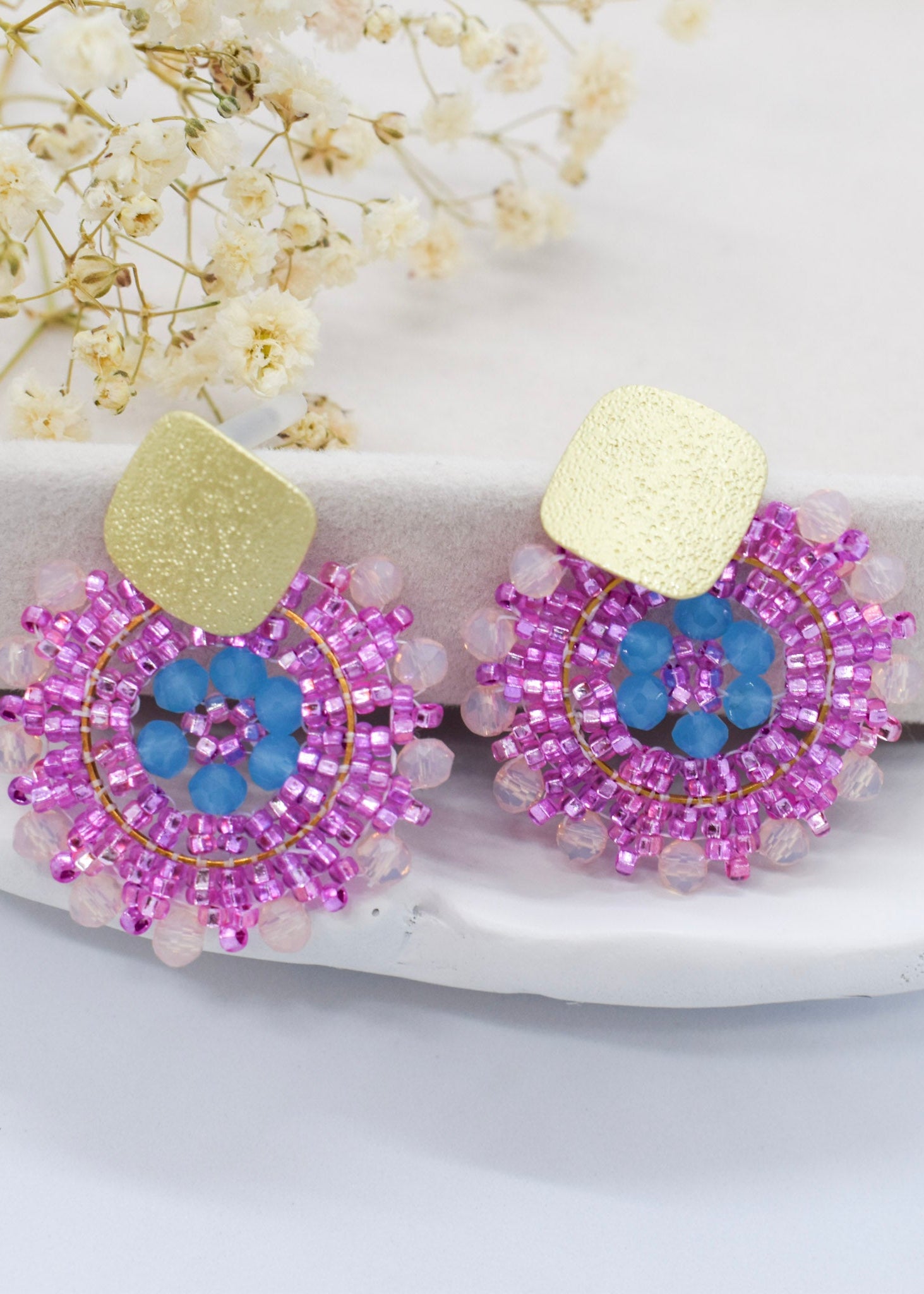 Pair of beaded earrings with pink and blue design on a white surface with flowers in the background