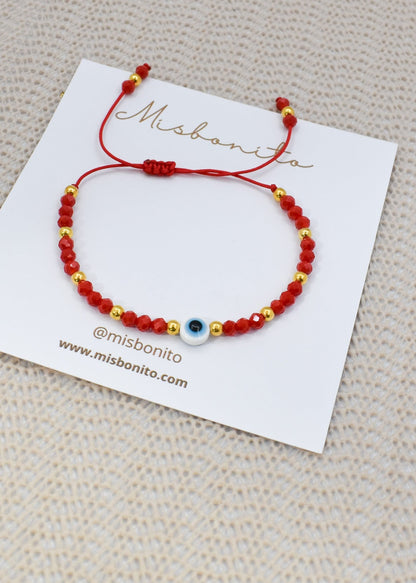 A red beaded bracelet with gold spacer beads and a central blue and white evil eye charm, displayed on a white Misbonito brand card over a textured beige fabric background.