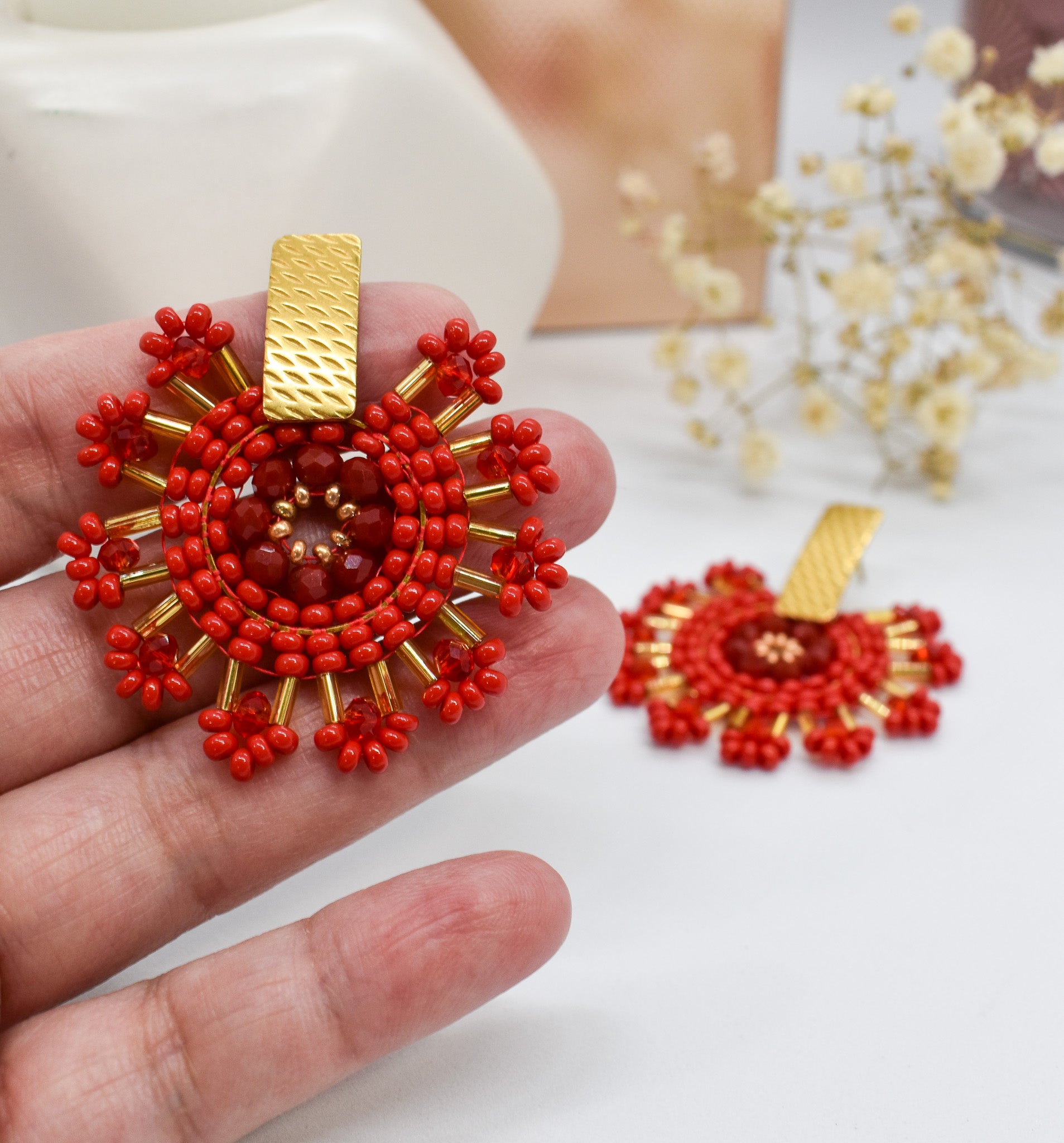 Red beaded earrings with gold accents held in a hand on a white background