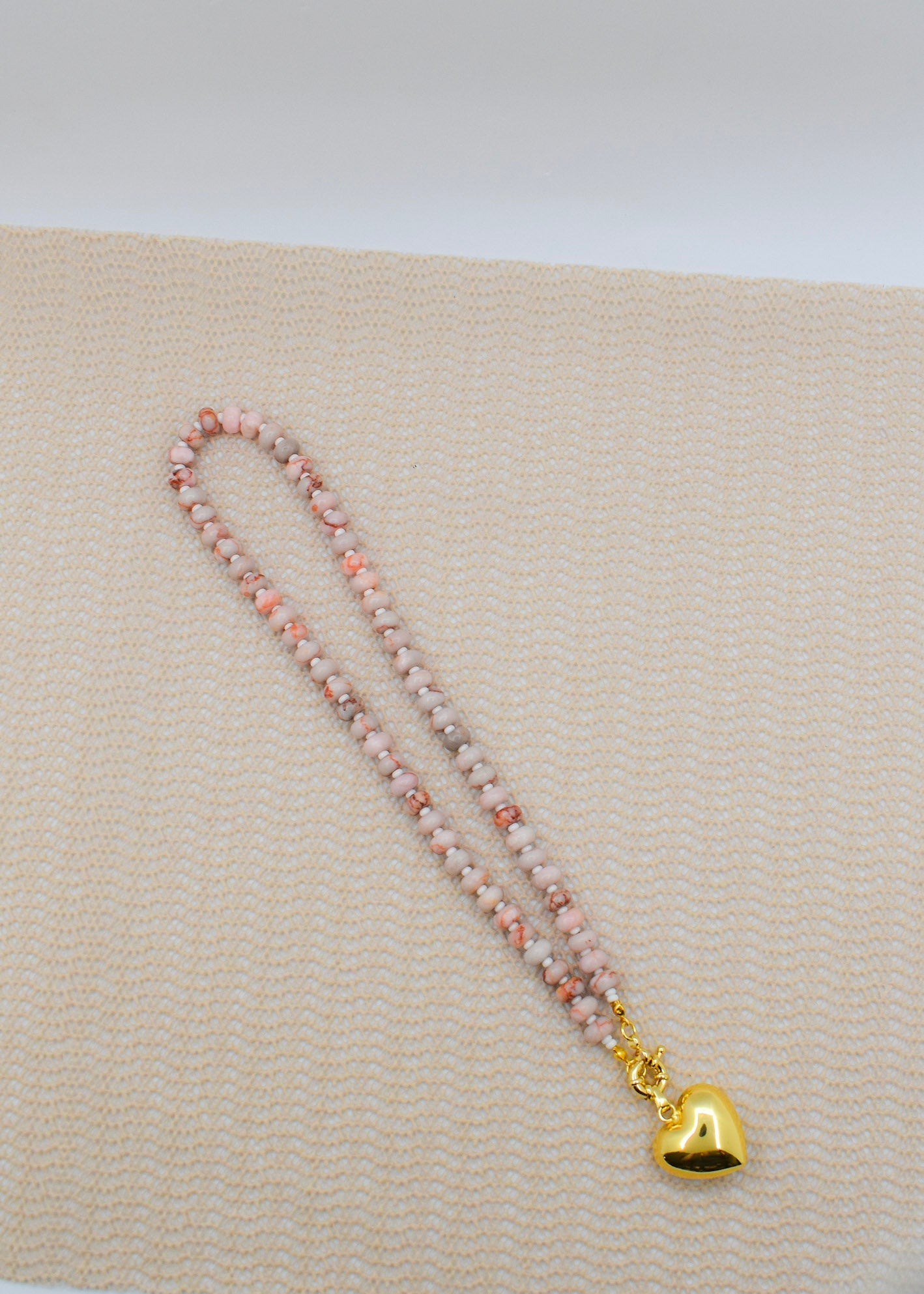 Necklace with pink beads and a gold heart charm on a beige fabric background