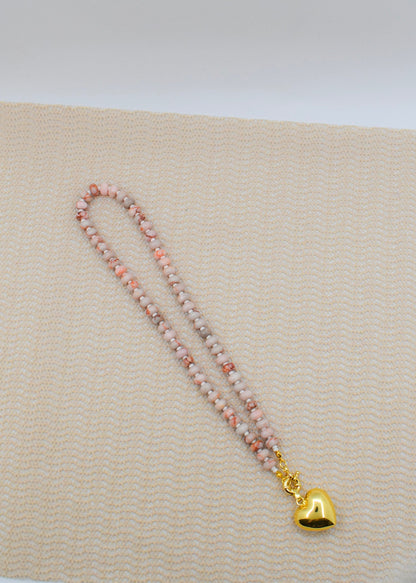 Necklace with pink beads and a gold heart charm on a beige fabric background