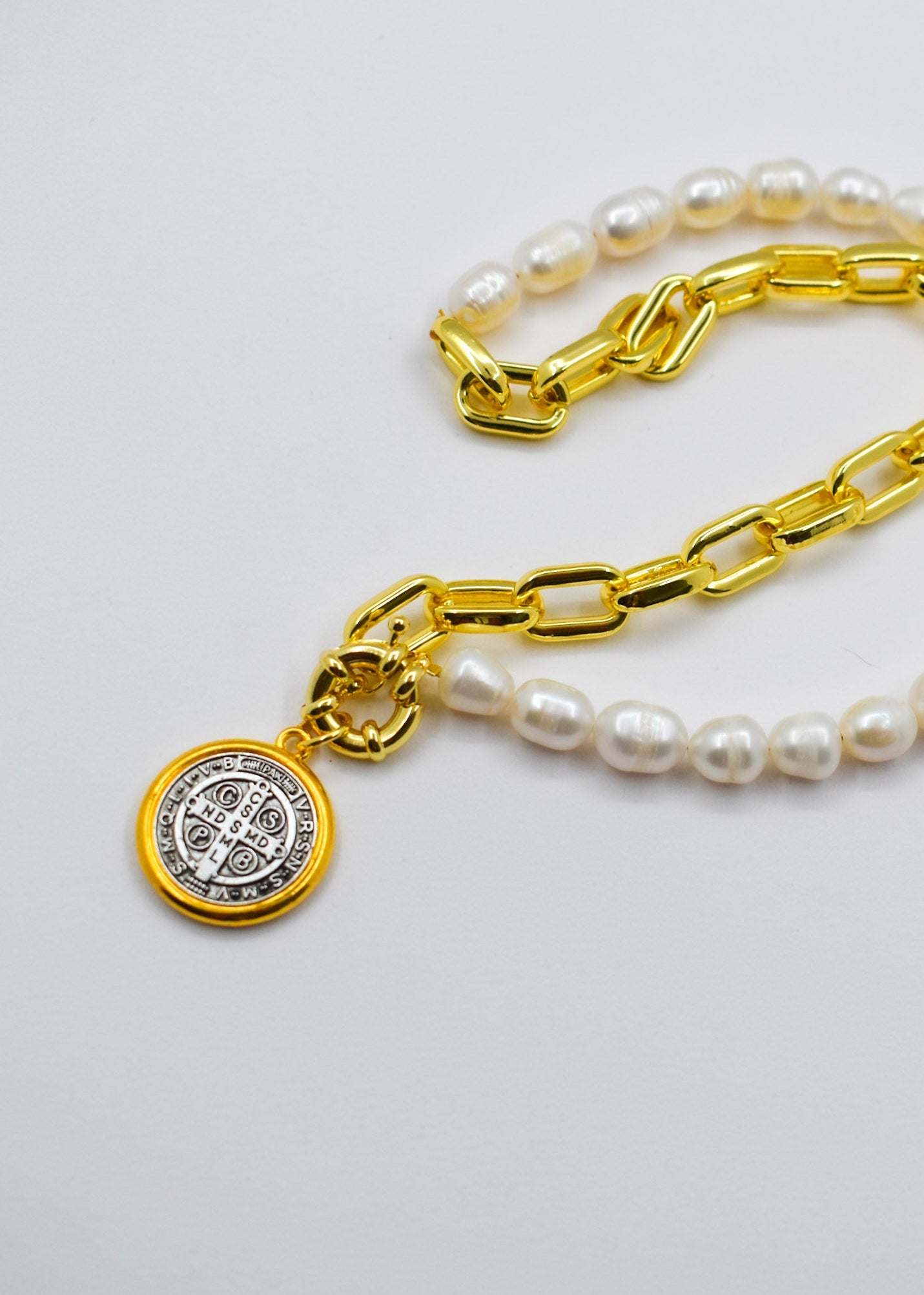A gold chain necklace with white pearl beads and a round gold Saint Benedict medal pendant, displayed on a light gray background.