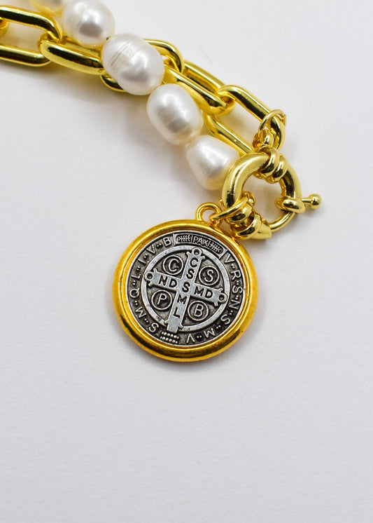 A close-up of a gold-tone chain bracelet with white pearl beads and a round gold-tone Saint Benedict medal charm, shown on a plain white background.