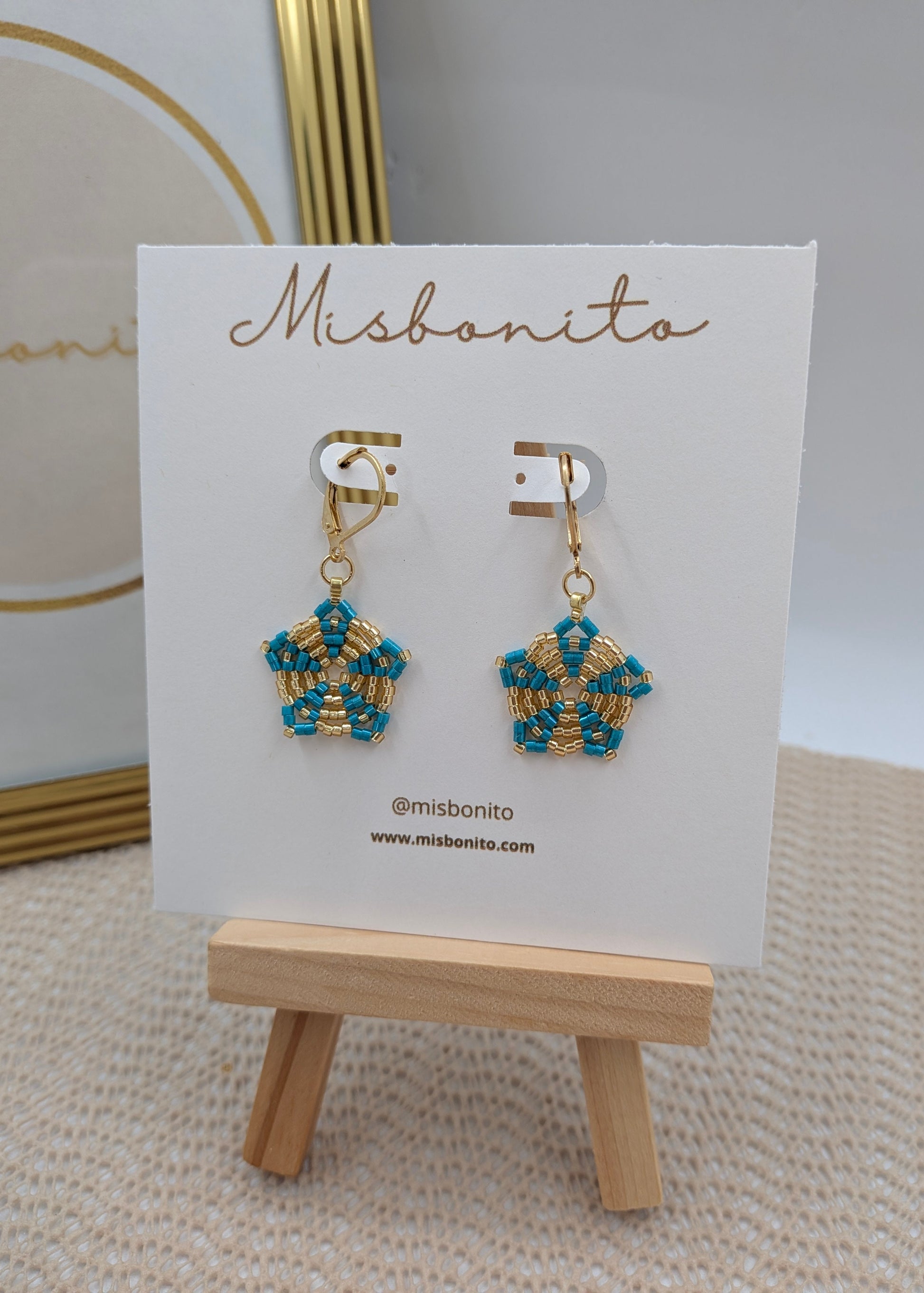 A pair of gold and turquoise beaded dangle earrings displayed on a white Misbonito card stand, which is placed on a small wooden easel.