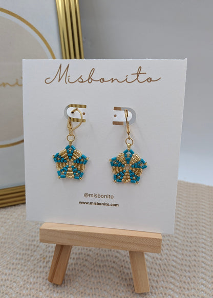 A pair of gold and turquoise beaded dangle earrings displayed on a white Misbonito card stand, which is placed on a small wooden easel.