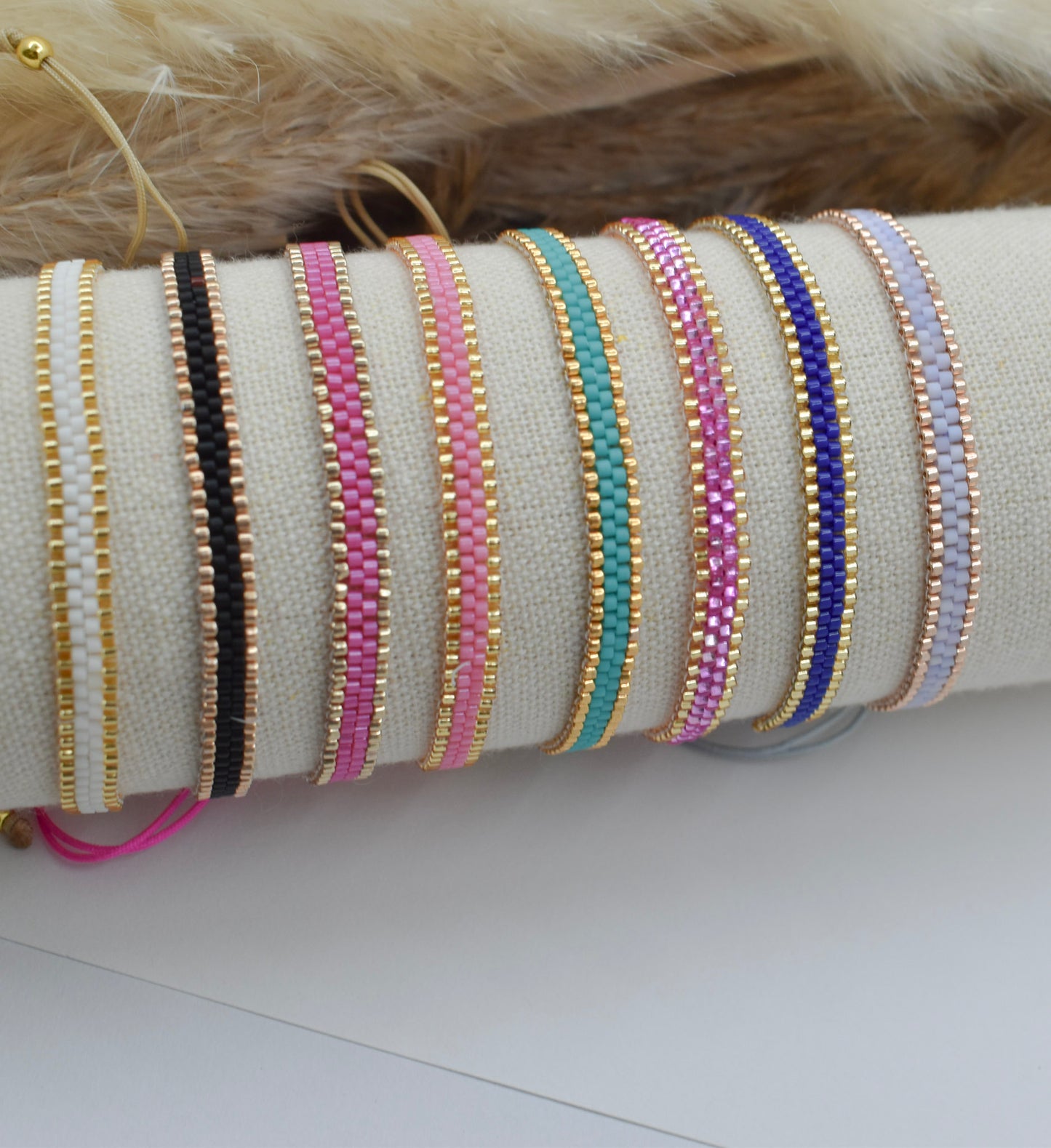 A set of beaded bracelets displayed on a cream fabric roll-up stand. The bracelets feature gold-plated metal links with seed beads in various colors including black, pink, mint green, purple, and light blue.