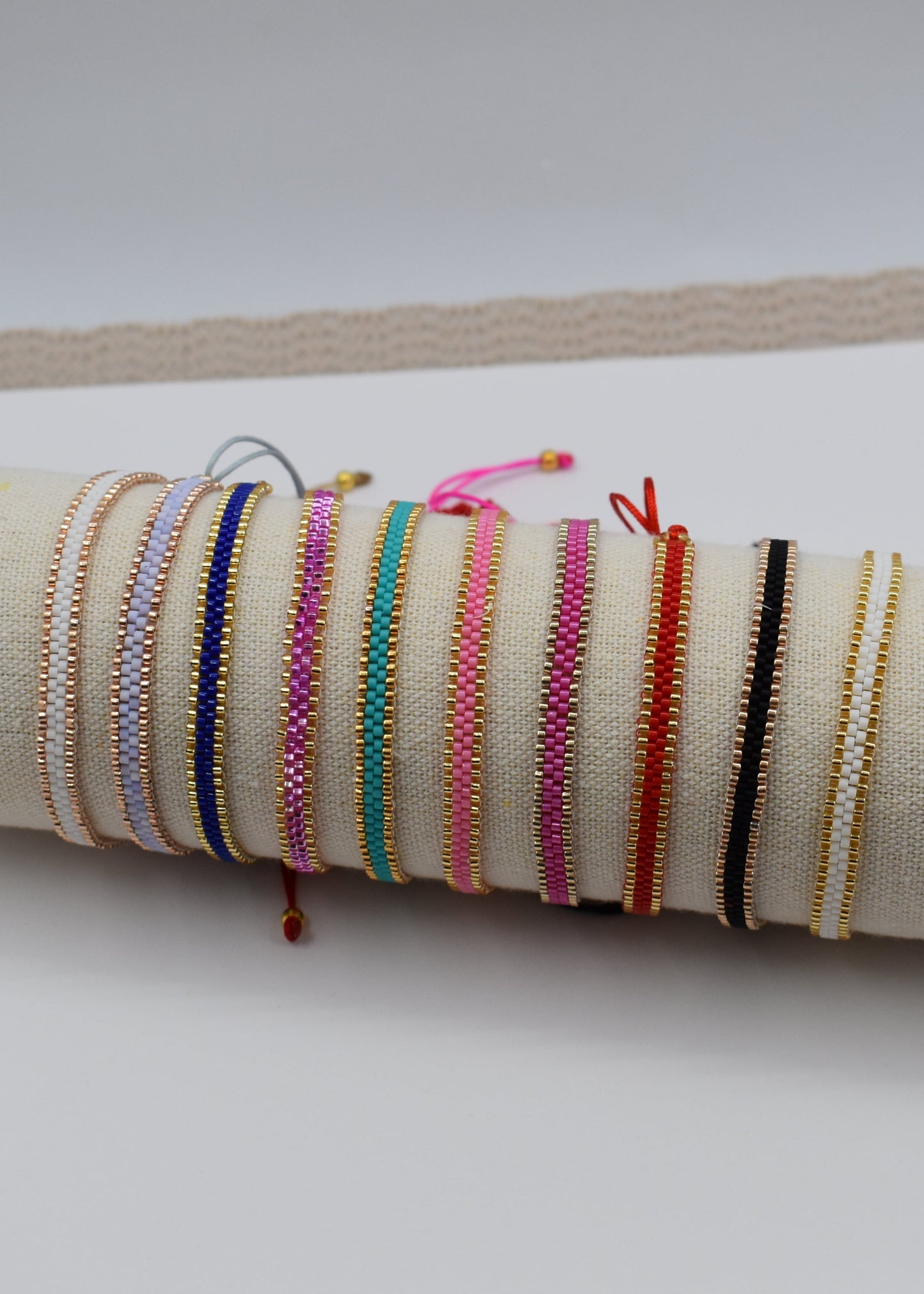 A collection of colorful woven cord bracelets displayed on a beige fabric-covered cylindrical stand, resting on a white card over a beige textured mat.