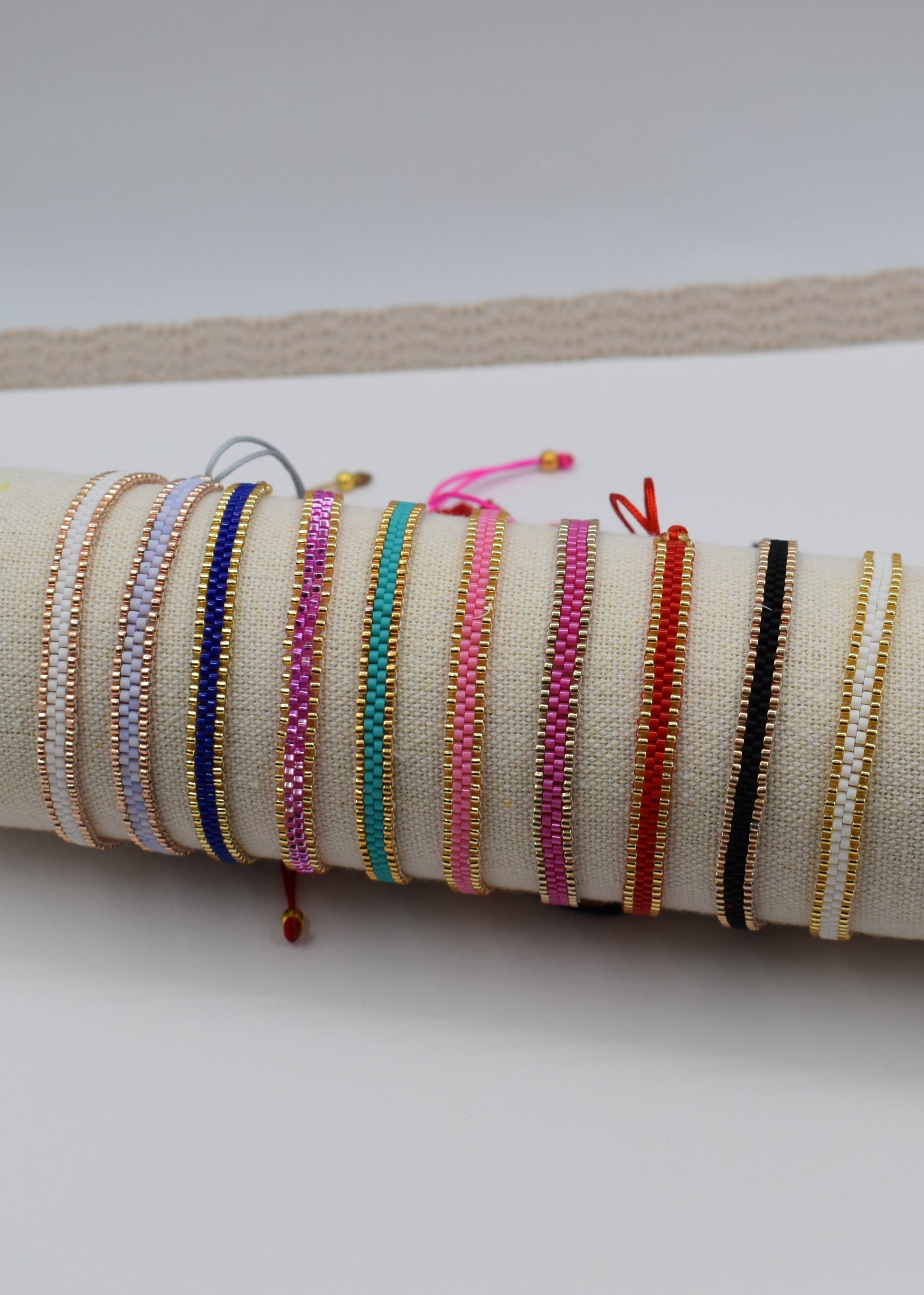 A collection of colorful woven cord bracelets displayed on a beige fabric-covered cylindrical stand, resting on a white card over a beige textured mat.