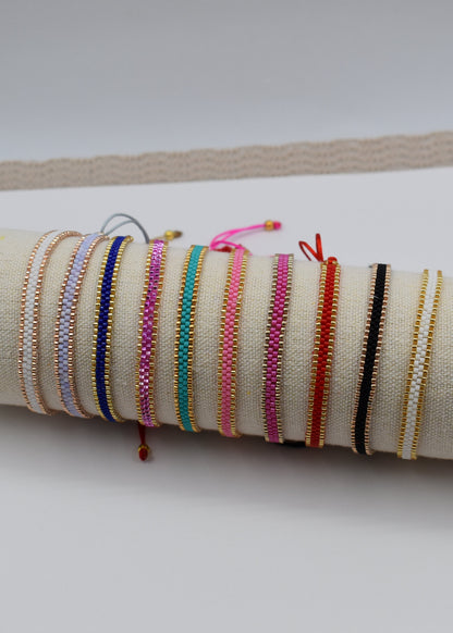A collection of colorful woven cord bracelets displayed on a beige fabric-covered cylindrical stand, resting on a white card over a beige textured mat.