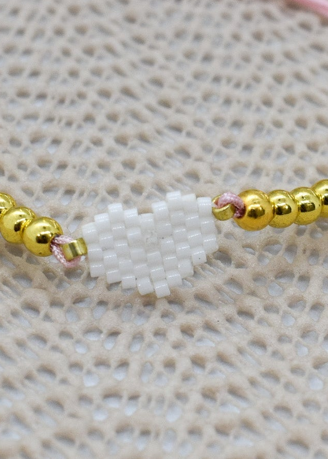 Zoomed Bracelet with gold beads and a white charm on a pink string against a textured beige background