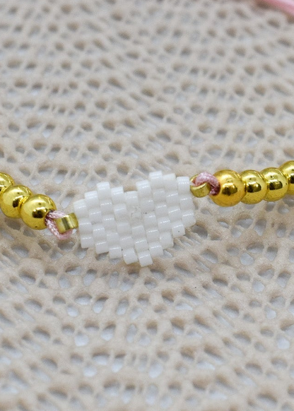 Zoomed Bracelet with gold beads and a white charm on a pink string against a textured beige background